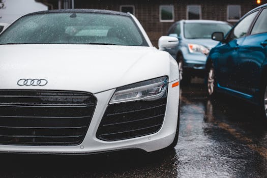 Modern white car with wet headlight and windshield parked on asphalt pavement near house